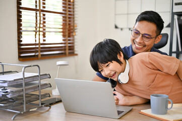 Happy Asian dad is being annoyed from his son while working in his home office.