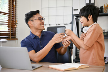 Adorable cute little son serving coffee to his father while he works in his home office.