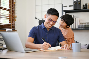 Happy Asian dad is being annoyed from his son while working in his home office.