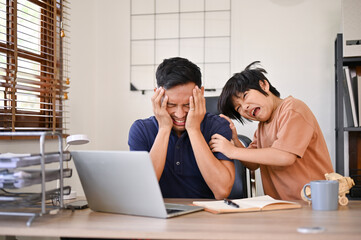 Unhappy Asian dad is being annoyed from his angry little son while working from home