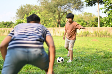 Cheerful young Asian boy playing football with his dad at the backyard, having fun time together
