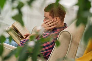 Homebound Tranquility. Man unwinding with a book in a cozy chair, amidst refreshing green plants at...