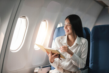Asian young woman take notes in a notebook sitting near windows at first class on airplane during flight,Traveling and Business concept