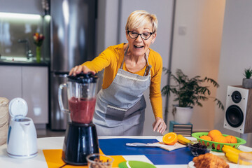 Senior woman in casual home clothes prepares healthy cocktails with different seasonal fruits.