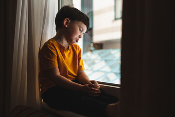 Portrait of sad and gloomy little boy sulking from disappointment, sitting on windowsill upset and...
