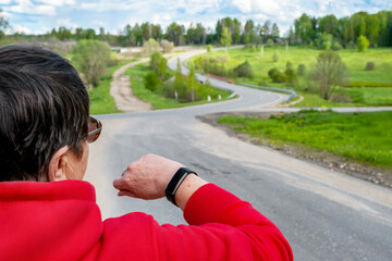 Unrecognizable elderly woman in red sportswear and glasses looks on screen of smartwatch outdoors. Concept of modern technologies and healthy lifestyle.