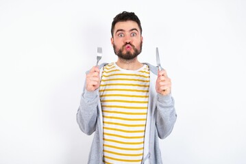hungry Young caucasian mán wearing trendy clothes over white background holding in hand fork knife...