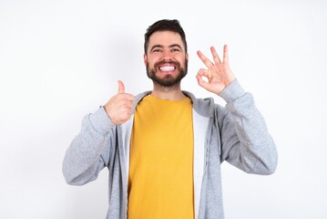 Young caucasian mán wearing trendy clothes over white background feeling happy, amazed, satisfied and surprised, showing okay and thumbs up gestures, smiling