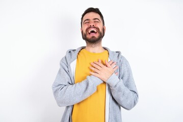 Young caucasian mán wearing trendy clothes over white background expresses happiness, laughs pleasantly, keeps hands on heart