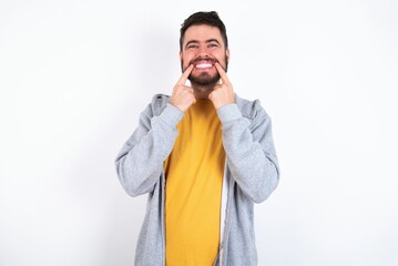 Fototapeta premium Happy Young caucasian mán wearing trendy clothes over white background with toothy smile, keeps index fingers near mouth, fingers pointing and forcing cheerful smile