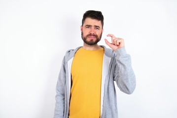 Displeased Young caucasian mán wearing trendy clothes over white background shapes little hand sign demonstrates something not very big. Body language concept.