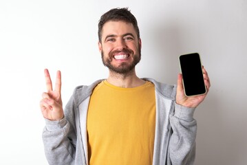 Young caucasian mán wearing trendy clothes over white background holding modern device showing v-sign