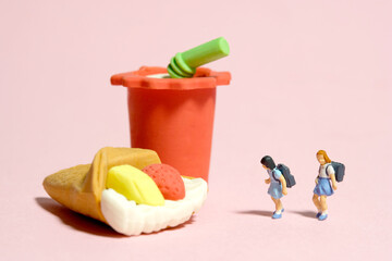 Miniature people toy figure photography. Healthy food eat and lunch break concept. Two girl kindergarten student standing in front of cake and soft drink. Isolated on pink background.