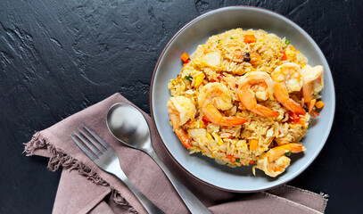 Fried rice with vegetables and sea prawns, carrots, eggs, and tomatoes. Thai Chinese food is arranged on a black plate on a black background kitchen table, top view.