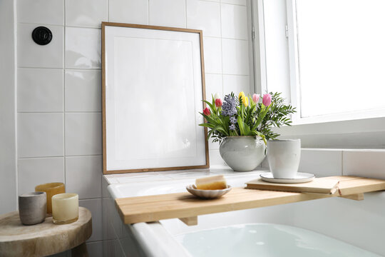 Elegant bathroom interior still life. Home spa relaxation concept. Bathtube with cup of coffee. Candles and book on board. Spring bouquet. Blank wooden picture frame mockup. White tiles background.