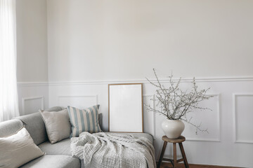 Spring home decor. Elegant scandinavian living room interior. Wooden picture frame, poster mockup on sofa. Linen pale blue striped cushions, throw. Cherry plum blossoms in vase, wooden stool.