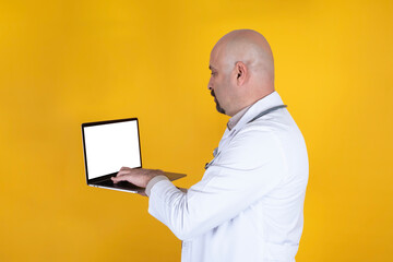 Doctor holding laptop, bald middle aged caucasian doctor holding laptop. White empty screen notebook mock up. Online treatment consulting, tele medicine concept idea. Isolated yellow background - obrazy, fototapety, plakaty
