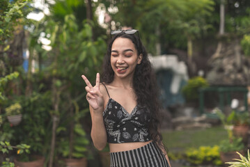 A playful woman makes a peace sign while sticking her tongue out. Outdoor scene at the garden.