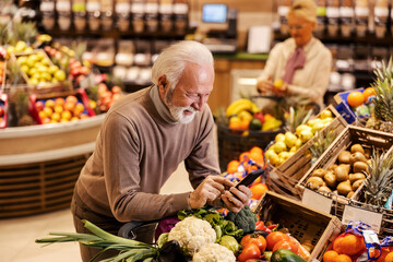 A senior man is leaning on shopping cart in supermarket and looking at the groceries list on cellphone.