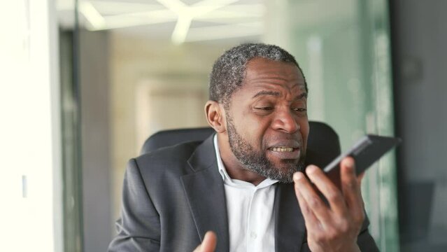 Close up. Angry mature african american man in formal suit talking and arguing on smartphone while sitting at workplace in modern office. Middle aged businessman shouting and proving problem solving
