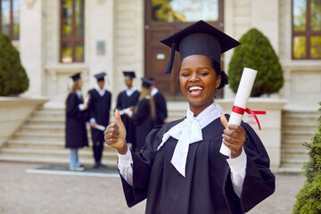 Portrait of happy cheerful joyful good successful beautiful African student girl in graduation cap and gown standing outside university building, holding diploma scroll, giving thumbs up and smiling