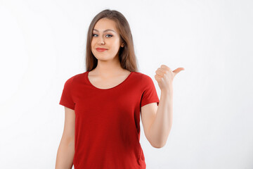 Fototapeta premium Happy lovely girl points finger at promo banner, shows advertisement on blank white space. Young brunette female stands over white background