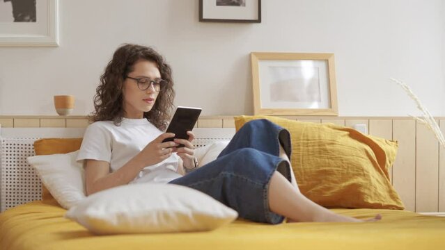 Pretty girl is reading a book in bright room at home. Beauty young woman enjoys reading and free time. Female student in glasses reads electronic book ebook in cozy bright living room, sitting on bed