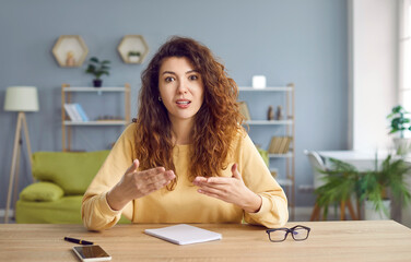 Portrait of teacher or coach at online conference or webinar. Young woman with beautiful long hair sitting at desk in front of camera, having video call, talking, explaining something, and gesturing