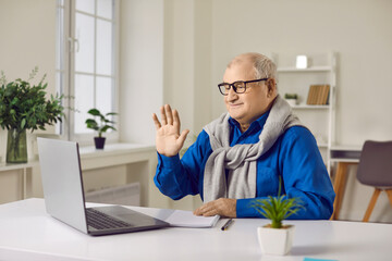 Happy old retired man waving hand talking to webcam making video call on laptop sitting at the desk at home. Gray -haired senior enjoying online communication technology looking at computer screen.