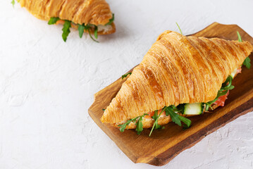 Crispy croissants sandwiches with salmon fish, cream cheese, cucumber and arugula on a wooden board.