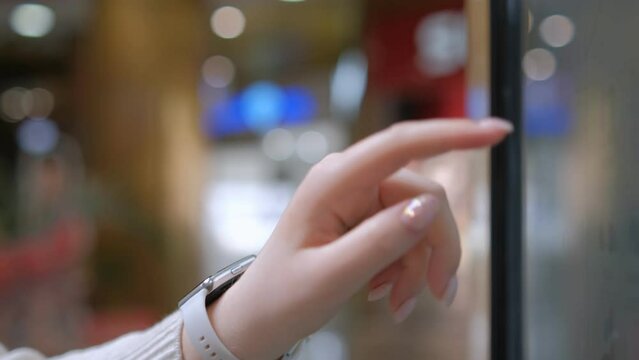 Woman using touchscreen terminal in mall. Detail view outstretched female hand touching modern big timetable or info screen in interior. Side view fingers swiping on display at out of focus background