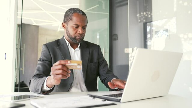 Happy african american man in formal suit typing credit card number on laptop while sitting at workplace in office. Businessman is shopping online and is satisfied with the good price of the product