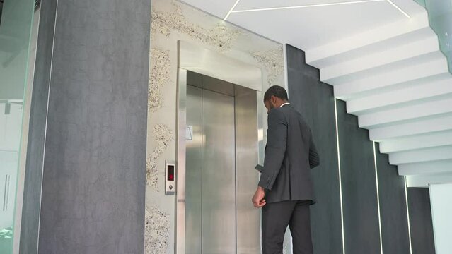 An african american businessman in a formal suit summons an elevator by pressing a button inside a modern office building. A middle aged male enters the lift elevator and goes upstairs to his work