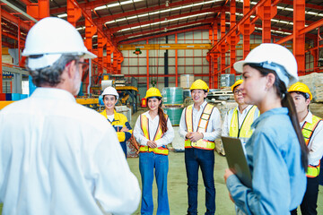 Group of industrial workers and engineers wearing safety helmets and vests attending team meeting in factory, representing workplace coordination and communication.