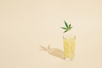 Sunlit glass of water with ice, lemon slices and marijuana leaf against bright sunny background. Minimal summer concept. Copy space.