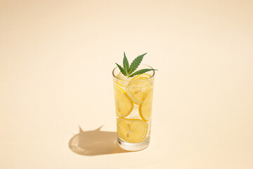 Glass of water with ice, lemon slices and marijuana leaf against bright sunny background. Minimal summer concept.