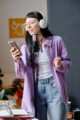 Young woman in headphones using her smartphone to sing song in microphone