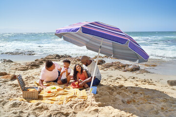 Gay male couple and kids eating lunch under umbrella on sunny beach