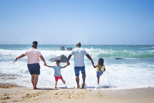 Gay male couple and kids holding hands, splashing in sunny ocean surf