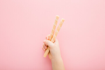 Baby girl hand holding two white vanilla wafers on light pink table background. Pastel color. Sweet snack. Closeup. Top down view.