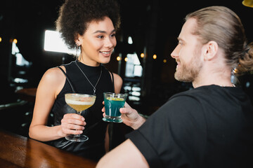 Smiling african amercan woman holding pisco sour cocktail near blurred friend in bar.