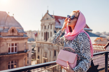 Outdoor portrait of young woman wearing retro scarf holding purse on balcony enjoying city architecture in spring