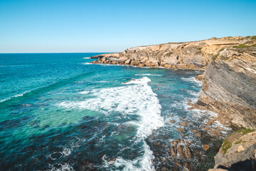 Breathtaking cliffs with crashing waves in the afternoon sun on the Atlantic coast near Vila Nova de Milfontes, Odemira, Portugal. In the footsteps of Rota Vicentina. Fisherman trail. Clear blue sky