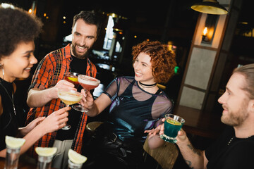 Overjoyed multiethnic friends holding glasses with cocktails near blurred tequila shots in bar.