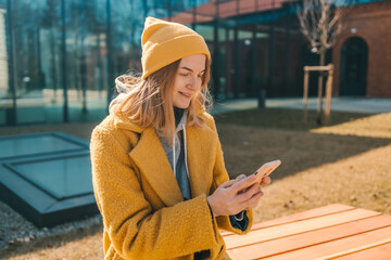 Fototapeta premium Young smiling happy student woman 30s wearing a yellow coat and hat sitting on bench in city spring park outdoors resting use mobile cell phone chat online. 