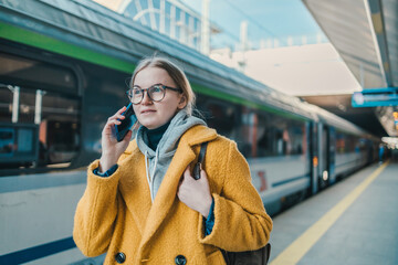 Fototapeta premium Young woman makes telephone call in roaming wears casual sweatshirt and yellow coat had glad expression waiting on a train station platform with passport and tickets enjoys smartphone talking. 