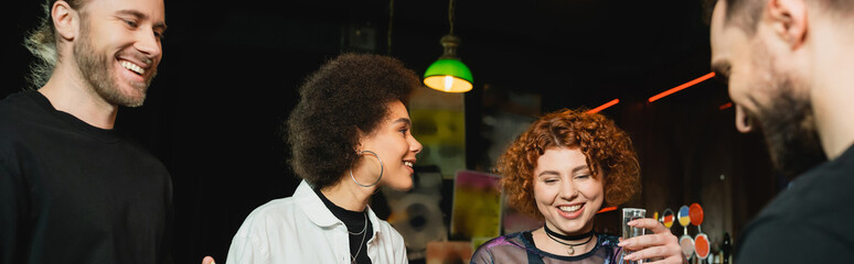 Smiling redhead woman holding tequila shot near multiethnic friends in bar, banner.
