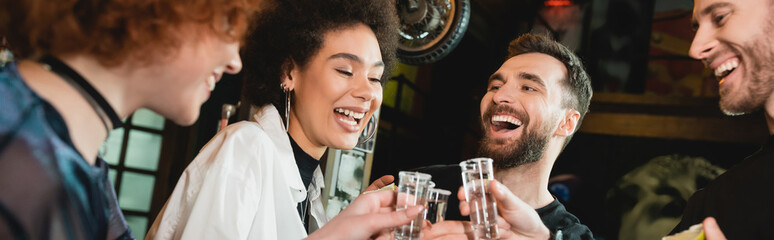 Cheerful interracial people holding tequila shots with salt in bar, banner.