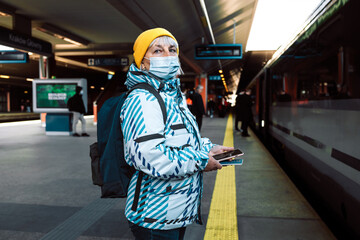 60s woman in face mask waiting on a train station platform with passport and tickets 