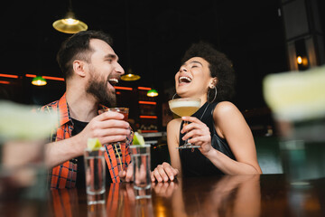 Laughing african american woman holding cocktail near friend and tequila shots in bar.
